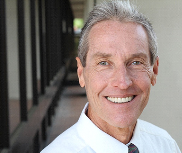 An older man wearing a button-down shirt and tie is smiling and showing off his new dentures in Carrollton