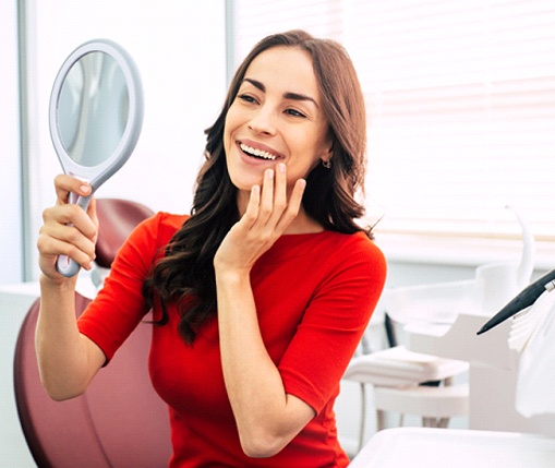 A young woman wearing a red blouse looks at her new smile in the mirror at a dentist’s office