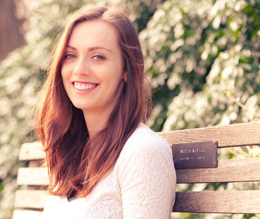 A young woman seated on a bench wears a white blouse and smiles, showing off her enlarged gums