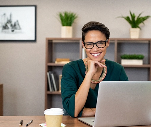 A woman sitting at a desk behind her laptop and smiling after receiving her metal-free dental crown